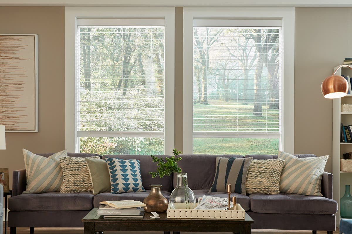 Living room pictured with big open windows that have white wood blinds letting sunlight pour through